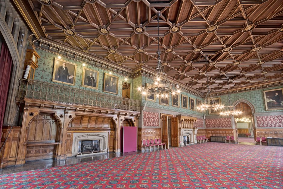 A large, well-lit banquet room with numerous round wooden dining tables arranged in rows, each decorated with small floral centrepieces in glass vases, set with white plates, silver cutlery, and drink