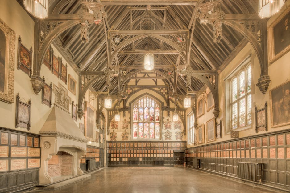 Interior of a large, historic hall with ornate wood paneling along the lower walls and decorative framed artwork hanging above. The ceiling features exposed wooden beams painted in a dark shade, with 