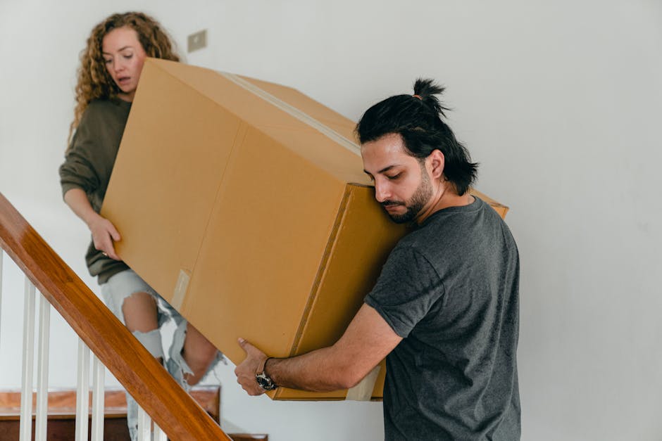 A man with dark hair tied in a small ponytail, dressed in a dark grey t-shirt, is carefully lifting a large cardboard box wrapped in clear packing tape while descending a staircase. Beside him, a woman with curly blonde hair, wearing a grey long-sleeved top and ripped denim jeans, is holding another similarly sized box and assisting with the move. The staircase has wooden handrails and white balusters, with a plain white wall in the background. The scene depicts a home relocation or furniture transport process within a residential property, emphasizing careful handling of boxed belongings during packing and moving activities. The lighting is natural, and the environment appears clean and orderly, supporting professional removal services such as those provided by Man with Van Cann Hall during house moves in Leyton maionette locations.