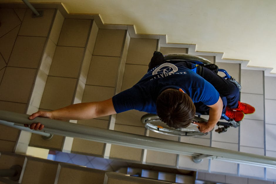 A man with dark hair tied in a small ponytail, dressed in a dark grey t-shirt, is carefully lifting a large cardboard box wrapped in clear packing tape while descending a staircase. Beside him, a woman with curly blonde hair, wearing a grey long-sleeved top and ripped denim jeans, is holding another similarly sized box and assisting with the move. The staircase has wooden handrails and white balusters, with a plain white wall in the background. The scene depicts a home relocation or furniture transport process within a residential property, emphasizing careful handling of boxed belongings during packing and moving activities. The lighting is natural, and the environment appears clean and orderly, supporting professional removal services such as those provided by Man with Van Cann Hall during house moves in Leyton maionette locations.