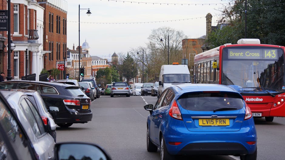 A busy street scene on Leyton High Road in Cann Hall, showing parked cars lining both sides of the road with a blue hatchback in the foreground, a red Metroline bus displaying route number 143 heading