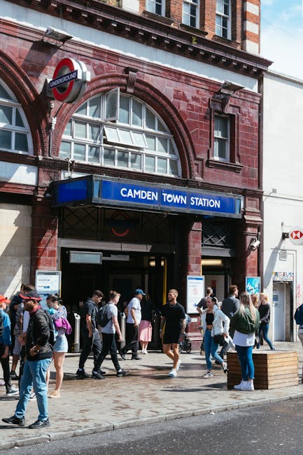 Outside Camden Town Station entry with a red-brick facade, large arched windows, and a blue sign indicating the station name. The underground roundel logo is visible above the entrance. In front, a diverse group of pedestrians, some carrying backpacks or shopping bags, are walking on the pavement near the entrance. The scene includes a wooden bench and a street scene with natural daylight. This setting relates to house removals and moving logistics by illustrating a common suburban or urban transit point near Leytonstone Station and Cann Hall, supporting the context of local relocations and furniture transport services provided by Man with Van Cann Hall, particularly during home relocation and packing and moving activities.