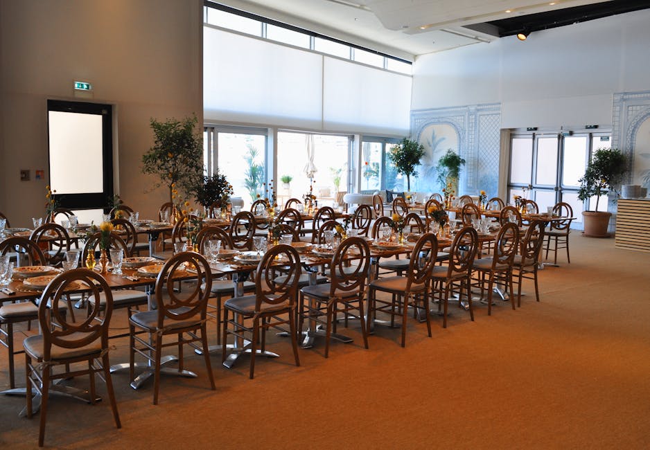 A spacious dining area with multiple tables set for an event, covered with white tablecloths and decorated with small floral centrepieces, situated in a well-lit room with large windows allowing natural light to fill the space. Wooden chairs with curved backs are arranged around the tables, some with fabric cushions. The room features high ceilings with modern lighting fixtures and a neutral colour palette, complemented by decorative blue wall panels and potted indoor plants placed near the windows. The floor is carpeted in a warm beige tone, and in the background, there are additional doorway entrances and a wooden counter or sideboard. This setting is indicative of an event space suitable for gatherings or formal functions, with the environment prepared for hosting guests. The image reflects a clean and organized interior, appropriate for professional moving and home relocation services such as those offered by Man with Van Cann Hall, focusing on packing and moving logistics in the context of house removals near Leytonstone Station and Cann Hall.