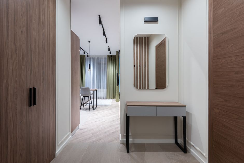 Inside a modern property showing the entryway with built-in wooden wardrobes on either side, featuring black handles. A small grey and wood console table with black legs is positioned beneath an oval mirror mounted on the white wall. In the background, the living area is visible, with a dining table and two chairs near a large window dressed with sheer white curtains and green drapes. The space is illuminated by ceiling-mounted black track lighting and pendant lights, with light-colored flooring. The image depicts a clean, well-organized interior during a home relocation process, potentially involving furniture transport and packing. The scene emphasizes the careful handling and organization of furniture and items typical of professional removals, as seen through the arrangement and environment managed by Man with Van Cann Hall.