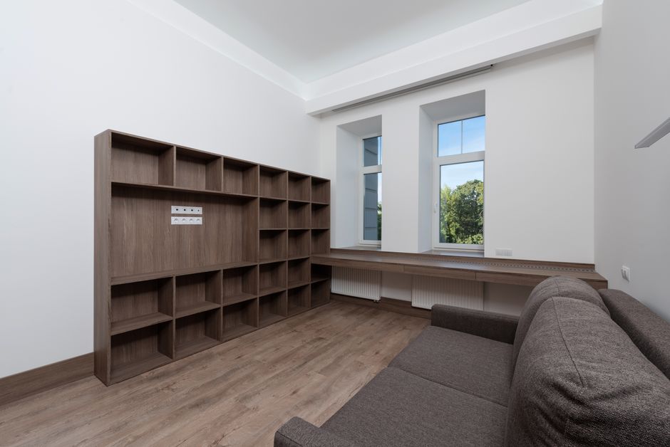 A spacious room with white walls and a light wooden floor, featuring a large empty wooden shelving unit with multiple open cubbyholes positioned against the left wall. To the right, a grey fabric sofa is partially visible, placed near the bottom corner of the image. The room has two tall, white-framed windows with clear glass panes, allowing natural daylight to illuminate the space and providing a view of greenery outside. Below the windows, a built-in wooden window seat with a radiator beneath extends across the width of the room. To the left of the window seat, a white electrical outlet is visible on the wall. The room appears tidy and well-maintained, suitable for a home relocation or furniture transport process, which is a typical part of packing and moving services provided by Man with Van Cann Hall. The overall environment suggests an interior space prepared for moving or storage, with clean lines and minimal furnishings.