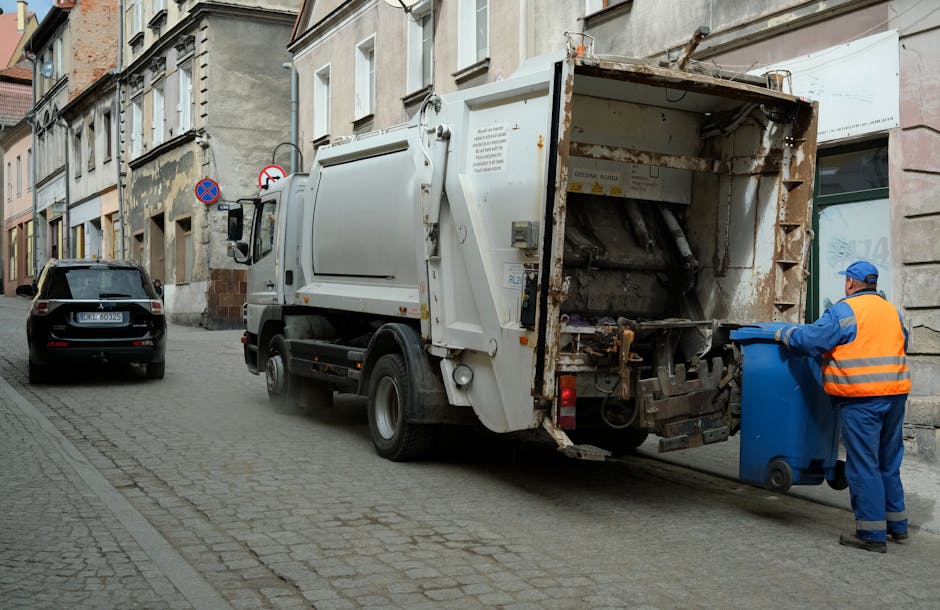A street scene in Cann Hall showing a waste collection vehicle operated by Man with Van Cann Hall, a white refuse truck with rusted areas near the rear loading area, positioned along a cobblestone street adjacent to residential buildings. A worker dressed in a blue uniform and reflective orange safety vest is placing a blue wheeled bin into the truck's rear compartment, which is open for waste collection. Several cardboard boxes and recycling materials are visible nearby, indicating ongoing home clearance or packing activities in the context of house removals or furniture transport. The scene is lit by natural daylight, with parked cars along the street, including a black car directly behind the truck, and a No Parking sign attached to the building on the left side. This image exemplifies the logistics involved in residential waste removal as part of property relocation and moving services offered by [COMPANY_NAME].