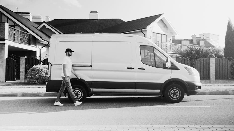 A man wearing a yellow and grey jacket, black gloves, and a cap stands next to a white cargo van parked on a paved driveway in a residential area. He is holding the van's door handle, preparing to loa