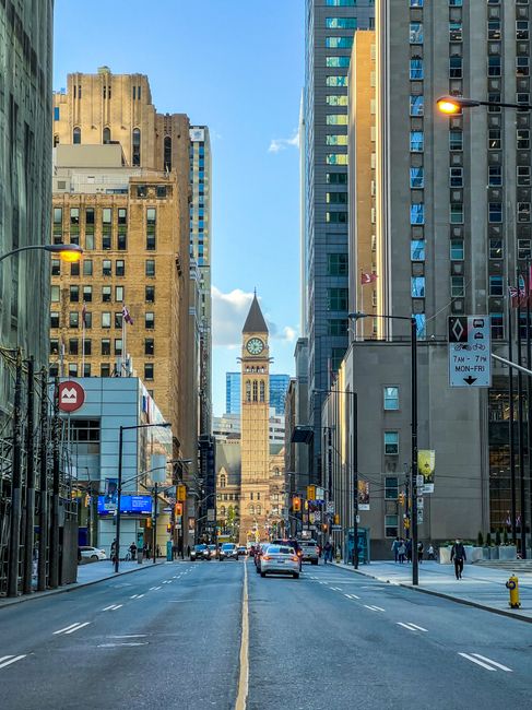 A wide urban street scene in Cann Hall during daylight, with high-rise office buildings and residential towers lining both sides of the road. In the distance, the iconic clock tower of the Royal Londons building is prominently visible against a clear blue sky with a few scattered clouds. The street features parked cars, moving vehicles, and pedestrians walking along wide sidewalks. Streetlights and signage are mounted on black metal poles, indicating parking regulations and traffic rules. In the foreground, a section of the road appears to be under preparation for a home relocation or moving process, with the presence of a small loading area on the pavement, possibly used by professional removals services like Man with Van Cann Hall for furniture transport or packing and moving activities. The overall scene captures an active city environment suitable for logistics and house removals, with clear visibility of urban architecture and street details.
