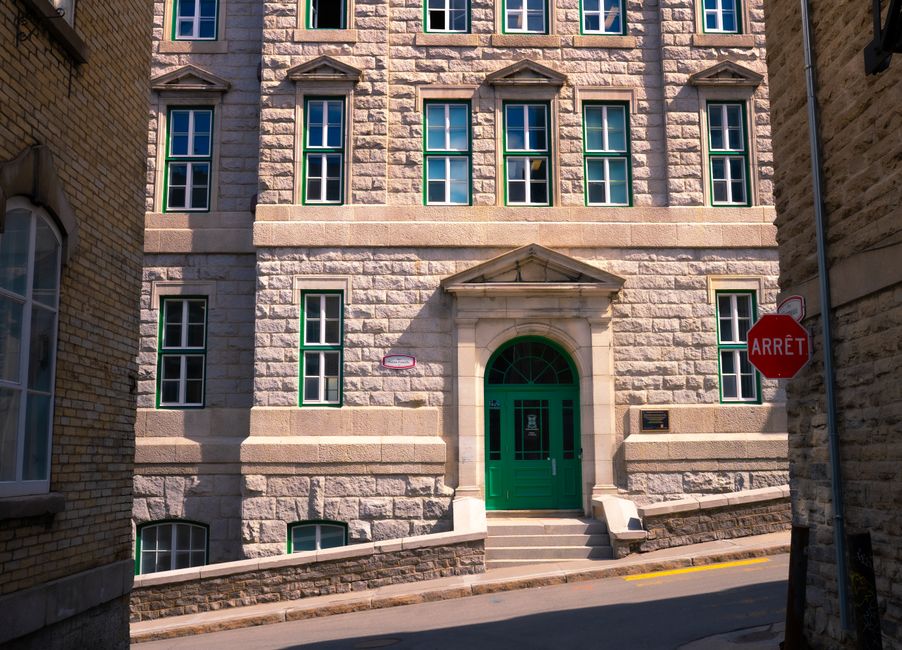 The image shows the facade of a historic stone building with multiple windows framed in white and green accents, including the main entrance which features a green door with a semi-circular transom window above it and a small stone pediment supported by triangular brackets. The building's exterior is constructed with large, rough-hewn stone blocks, and a concrete steps lead up to the doorway. To the right of the entrance, there is a red stop sign with the word 'ARRÊT' mounted on a metal pole on the narrow street. The sidewalk in front of the building is bordered by a curb, and there is a small plaque and a red mailbox affixed to the wall near the door. The image is well-lit with natural daylight, highlighting the architectural details and the texture of the stonework, set within an urban environment that suggests a well-preserved historic area. This scene is relevant to moving and relocation services provided by Man with Van Cann Hall, including considerations for street permits during home relocation projects.