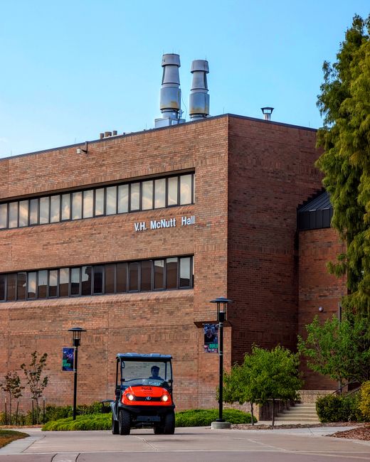 Exterior view of V.H. McNutt Hall, a brick-built university building with three large ventilation stacks on the roof, situated in daytime with clear blue sky. In the foreground, a small red and black utility vehicle is parked on a paved area near the entrance, surrounded by neatly landscaped greenery and young trees. Two lamp posts with banners stand along the paved path leading to the building's steps. The scene depicts a campus environment during a home relocation or moving process, with no people visible, emphasizing the building's facade and surrounding outdoor space. Man with Van Cann Hall specializes in removals and moving services, often involving loading and transport of furniture and boxes through this type of setting.