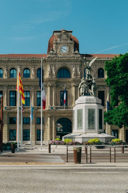 Photo of a historic municipal building with a central clock tower topped by a red, domed roof, set against a clear blue sky. In front of the building, there is a white stone monument featuring multiple sculptures, including a seated figure and a winged figure, mounted on a rectangular pedestal with inscriptions. The area surrounding the monument is paved with concrete, with several flagpoles displaying various national and regional flags positioned along the front of the building. To the right, there are green trees providing partial shade, and a metal railing separates the pedestrian area from the street. The scene is well-lit, with natural sunlight highlighting the architectural details of the building and providing a neutral, factual context suitable for an article about house removals or relocation services, subtly supporting themes of urban moving logistics.