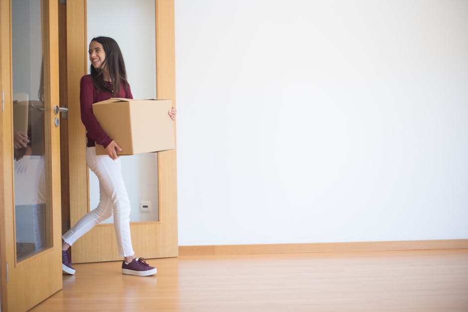 A young woman with long dark hair smiling and holding a medium-sized cardboard box, dressed in a maroon top, white pants, and maroon sneakers, stands in a room with light wooden flooring and a white wall. She is partially outside a wooden-framed glass door, which is open. The door features a glass panel with a wooden handle and a hinge visible on the left side. In the background, there is minimal furniture or decoration, indicating an empty property ready for moving. The woman appears to be in the process of packing or moving items, part of a home relocation or furniture transport scenario. The setting is well-lit with natural light, emphasizing the clean and uncluttered environment. This scene naturally aligns with professional removals and packing services provided by Man with Van Cann Hall, supporting house removals for flats on Cann Hall Road, Leyton E10.