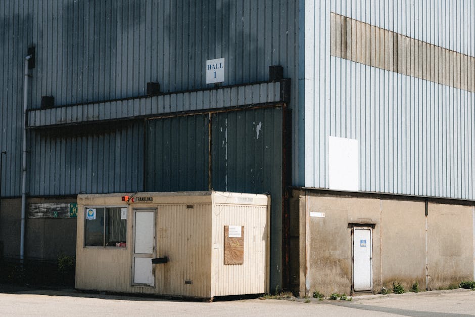 Exterior view of a large industrial warehouse in Cann Hall, Leyton E10, featuring corrugated metal siding painted in light blue with a horizontal beige stripe near the top. In front of the building is a beige, portable security cabin with a white door, which appears used as a site office or storage, with some signs and notices affixed to its surface. The cabin is positioned on the pavement adjacent to a large sliding metal loading bay door, painted in darker blue, with signs of rust and wear. On the right side of the image, a smaller, plain white door is embedded in the warehouse’s concrete lower section. The area around the cabin and warehouse is well-lit with direct sunlight, casting shadows on the ground, and the scene depicts a typical setting for loading and unloading furniture and relocation supplies during a house or flat move. Man with Van Cann Hall may coordinate such moving logistics involving this kind of warehouse environment for home relocations and furniture transport.