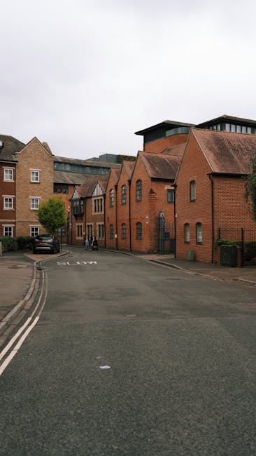 A quiet street scene on Cann Hall Estate featuring a narrow residential road with a gentle curve, lined with brick buildings including houses and small commercial or industrial-style structures. The buildings exhibit varied roof shapes, including gabled and flat roofs, with some windows visible on their facades. Several parked cars are along the curb, and a few pedestrians can be seen further down the street near the buildings. The scene is captured in natural daylight under overcast skies, with muted lighting. The image shows no active loading or moving operations, but it sets a context for house removals, highlighting the typical urban environment where furniture transport and home relocation services by Man with Van Cann Hall could take place. The environment appears organized and accessible for vehicle parking and moving activities in line with the company's removals services.